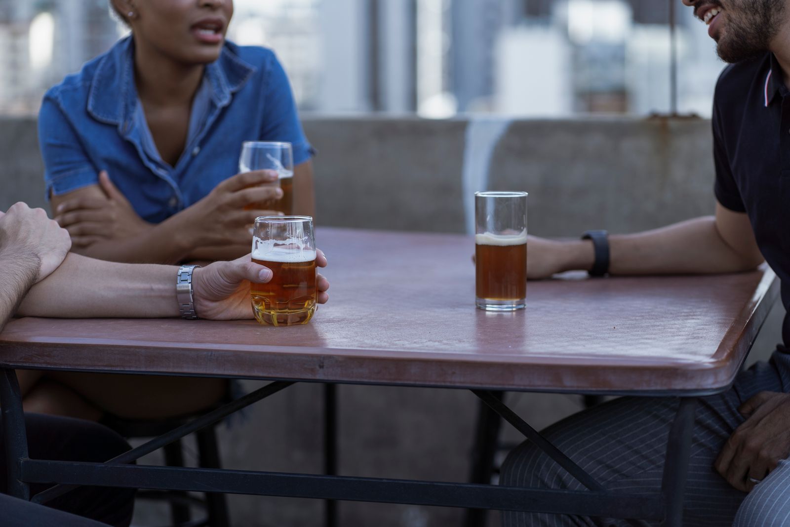 Three plain glasses of amber beer on a rust-red metal outdoor table with two people mid-conversation, rooftops blurred behind in early-evening light.