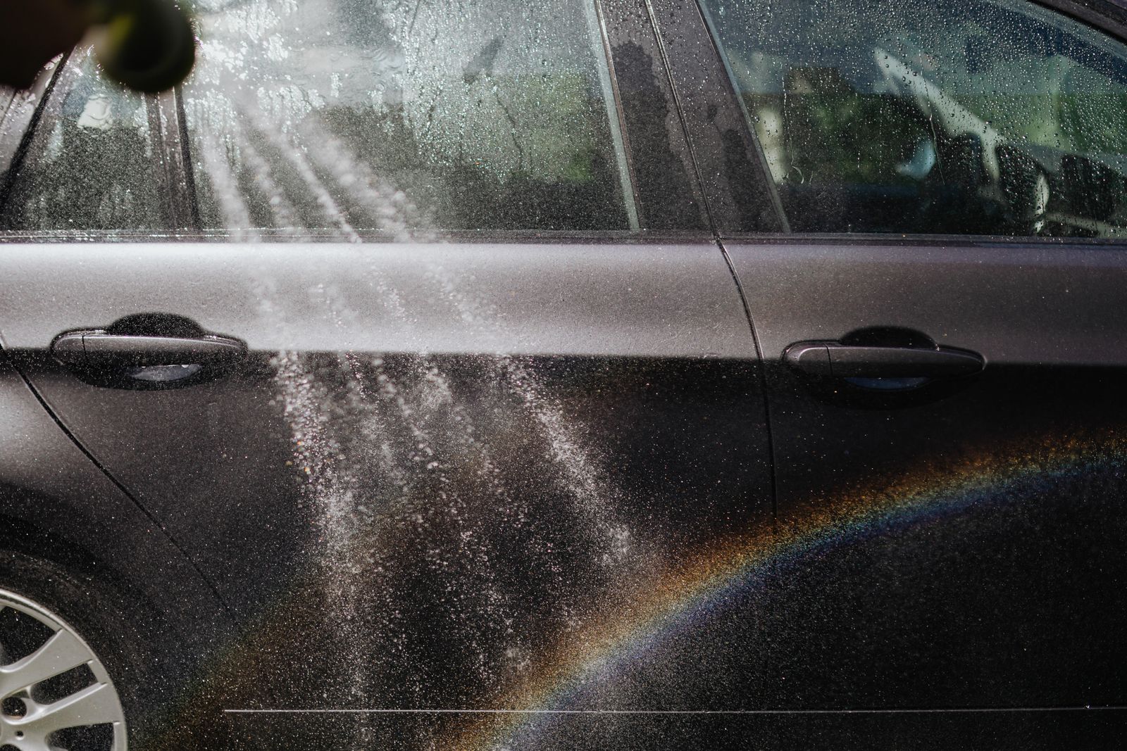 The side of a dark car being rinsed with a fine spray of water in bright sunlight, the spray catching the light.