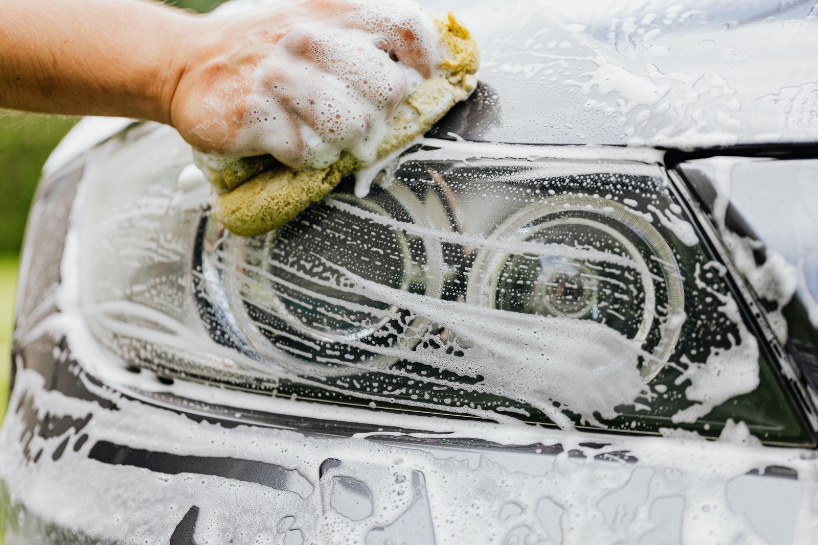A bare hand holding a yellow sponge thick with white soap suds, scrubbing the headlight and bonnet edge of a silver car, with blurred green foliage behind.