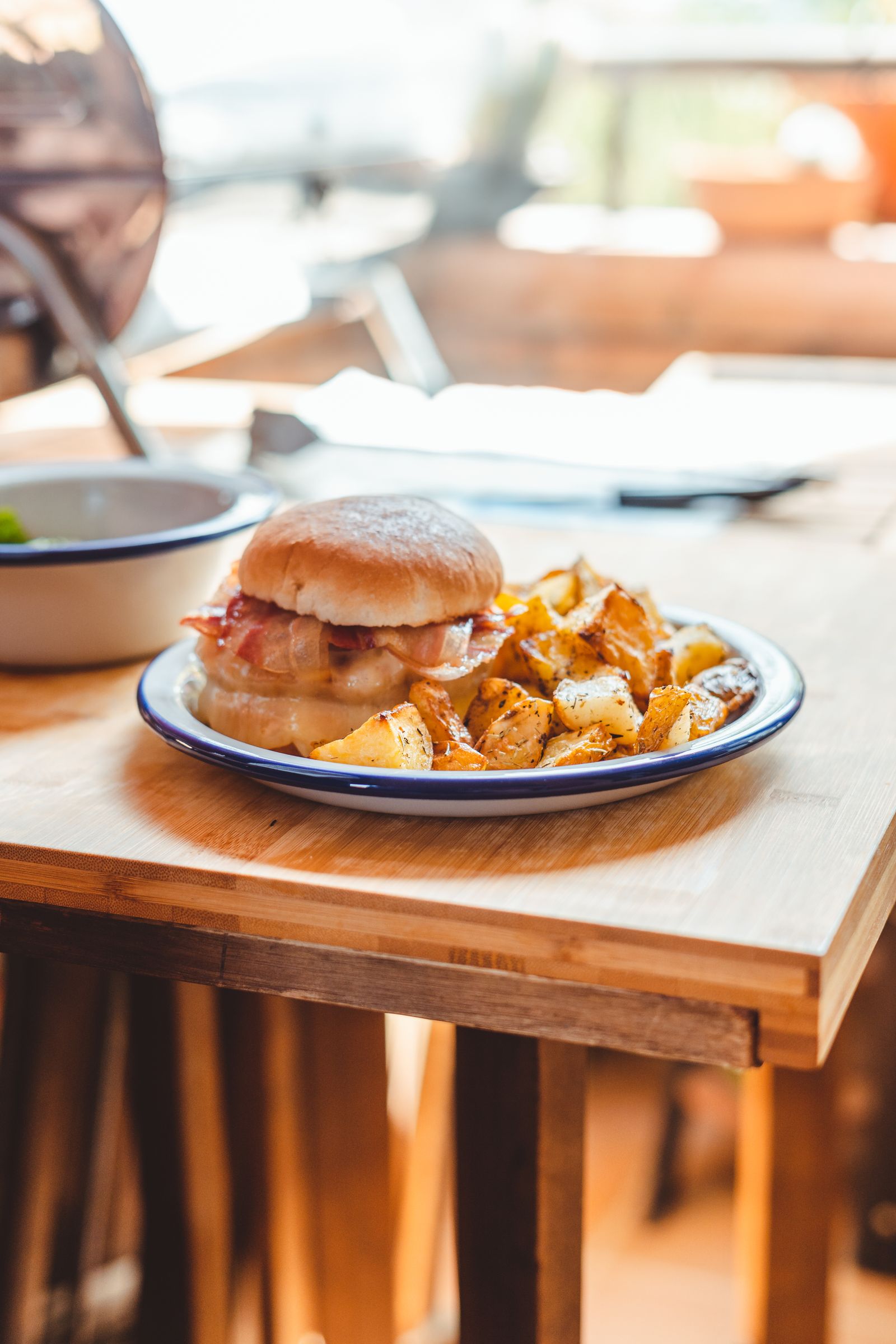 A bacon burger with golden roast potato chunks on a white enamel plate with a dark blue rim, on a light wooden table in warm daylight.