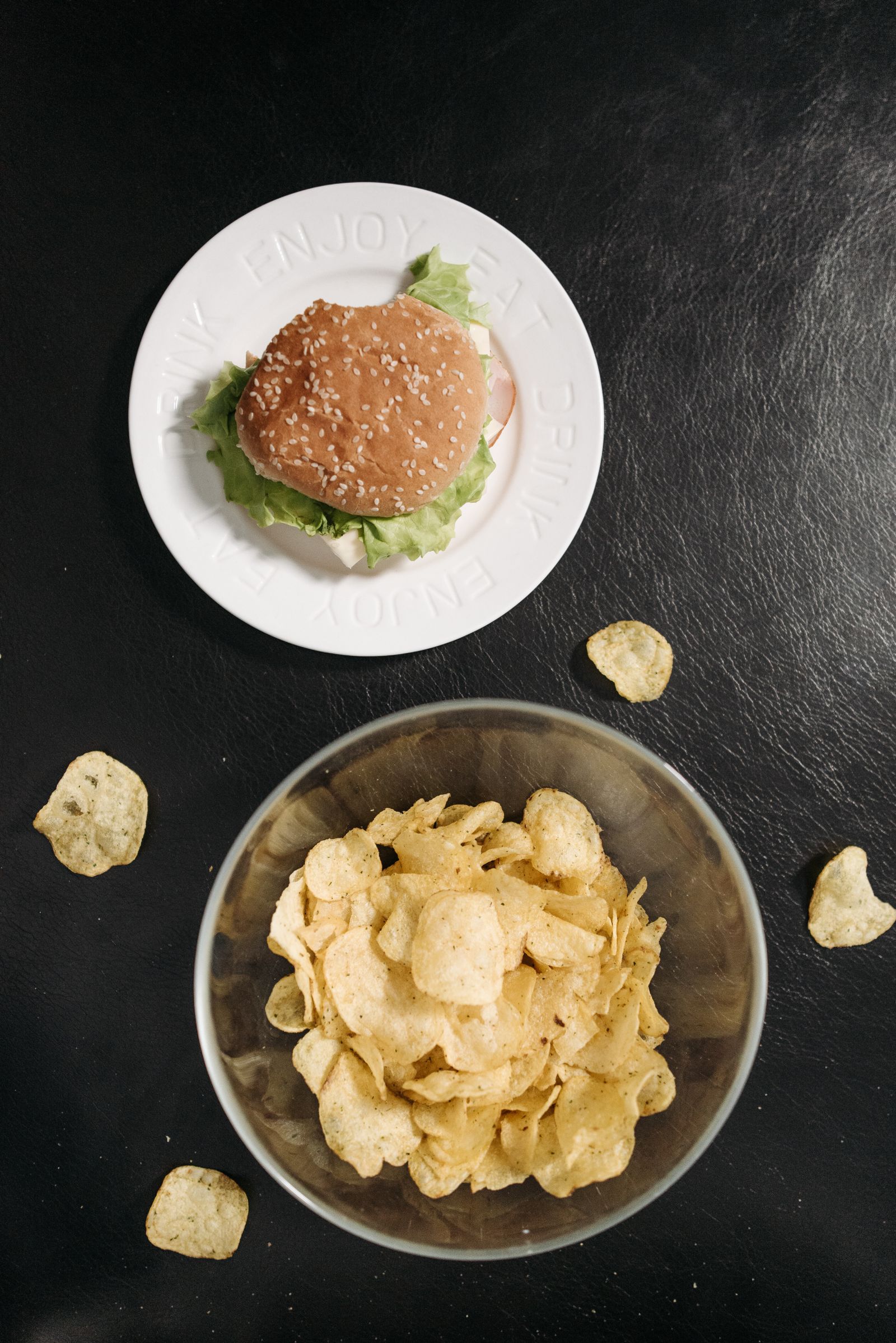 Top-down view of a burger in a sesame-seed bun with lettuce on a round white plate next to a metal bowl heaped with rustic potato crisps, a few loose crisps on a dark surface.