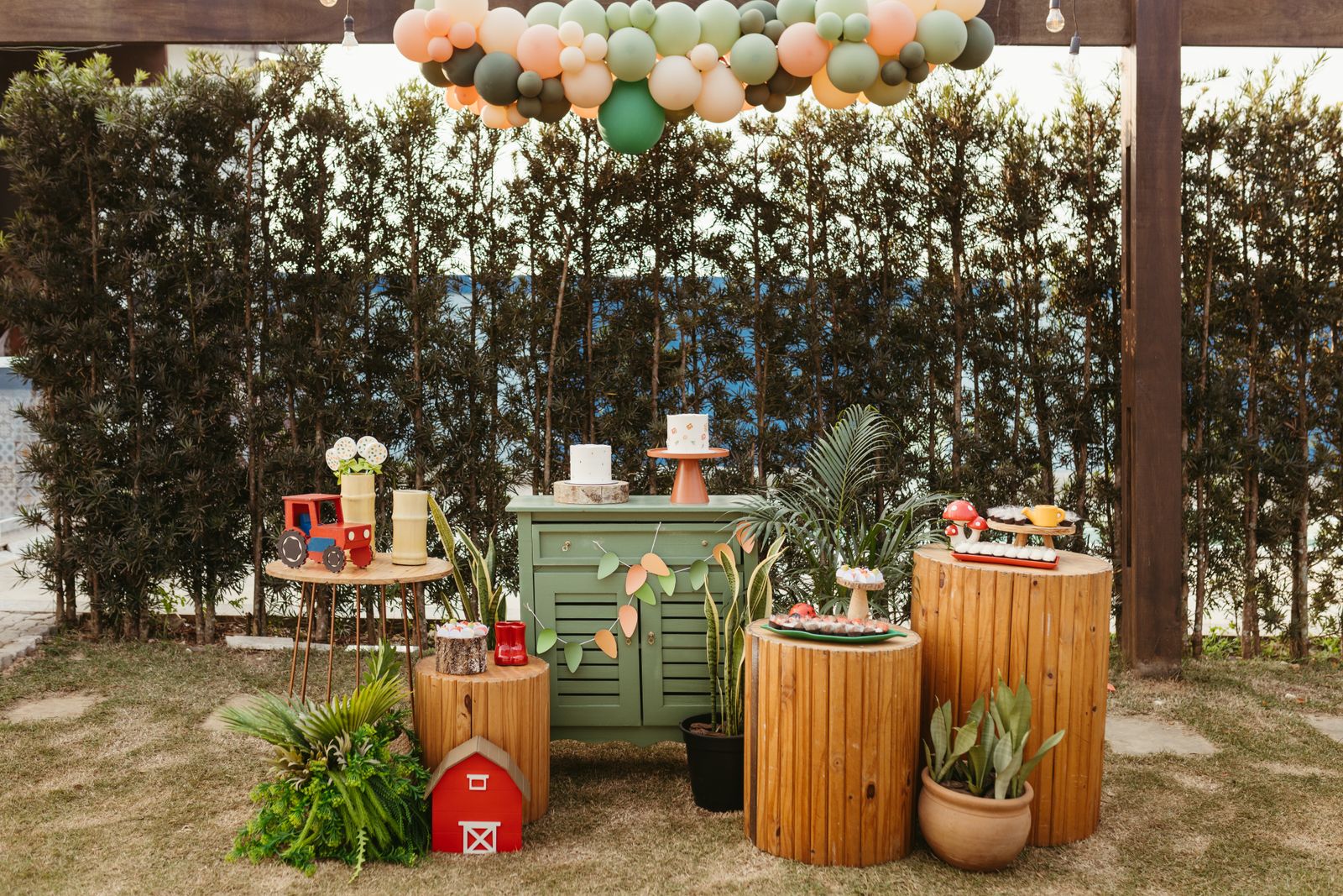 An outdoor kids' party setup on a grass lawn against a tall green hedge under festoon lights: a sage-green, peach and cream balloon garland, a cabinet with a small two-tier cake, wooden log-slice plinths with trays of treats, potted plants and toy tractor props.